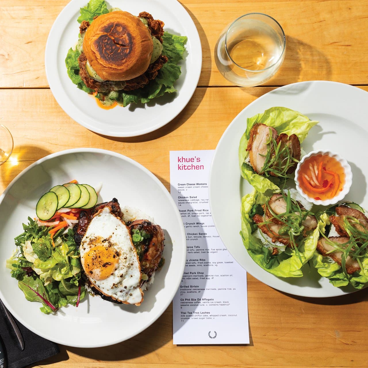 Three plated dishes from Khue's Kitchen displayed on a wooden table: a Spicy Chicken Sandwich on a bun with lettuce, Crispy Pork Lettuce Wraps with dipping sauce, and a Grilled Thick-Cut Pork Chop topped with a fried egg and served with a fresh salad. A menu and glasses of wine are also on the table.