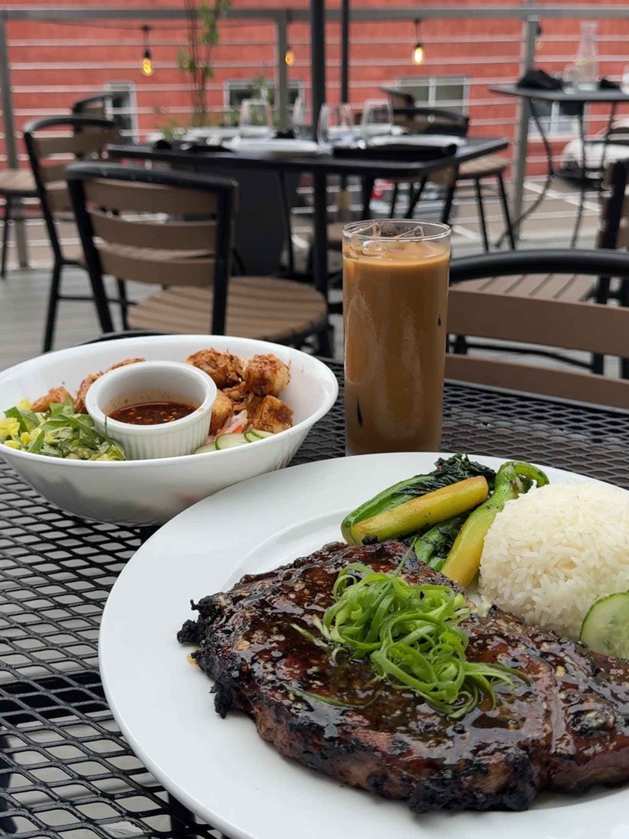 Close-up of our Grilled Ribeye, next to a bowl of Bún Chay and a tall glass of Cà Phê Sữa Đá Coffee, set on our outdoor patio with tables and chairs in the background.